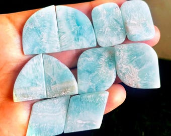 May include: A collection of polished larimar gemstones in various shapes, including half-circles, rectangles, and rounded squares. The stones display a light blue color with white and darker blue patterns, resembling ocean waves. The gemstones are held in a hand.
