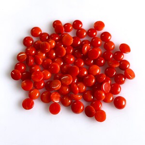 May include: A collection of numerous small, round, smooth, and polished red coral beads. Each bead features a vibrant, uniform orange-red hue with a glossy finish, arranged in a loose, scattered pile against a plain white background.