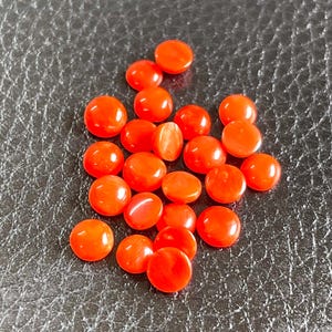 May include: A collection of small, round, bright orange cabochons with a smooth, polished surface. Each piece features a domed top and a flat base, exhibiting a vibrant, solid color throughout with subtle light reflections on the curved surfaces.
