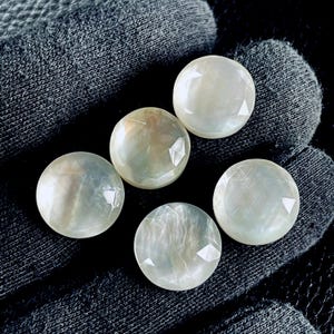 以下が含まれることがあります： ファセット加工されたエッジを持つ、5つのイリデッセントな丸い宝石。宝石は真珠のような光沢があり、微妙な色の遊びで光を反射します。石は濃い灰色のテクスチャのある表面に配置されています。