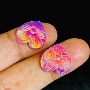 May include: Two heart-shaped cabochons featuring a carved floral pattern. These translucent pieces exhibit a vibrant iridescent finish with shifting hues of pink, purple, and yellow, reflecting light across their smooth, rounded surfaces.