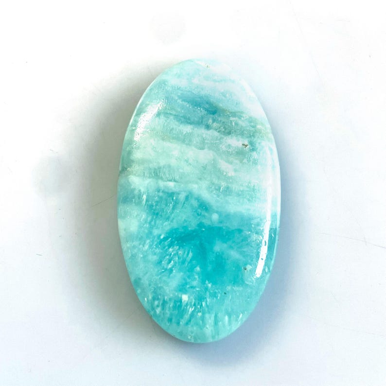 Peut inclure: Une pierre pr&eacute;cieuse polie de forme ovale pr&eacute;sentant une surface lisse avec un d&eacute;grad&eacute; de couleurs allant du turquoise profond et du cyan vif au blanc doux, affichant des motifs cristallins naturels et des inclusions min&eacute;rales.
