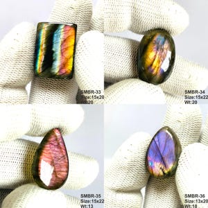 May include: Four polished labradorite gemstones in various shapes. The stones display iridescent colors including blue, purple, yellow, and pink. Each stone is held by a gloved hand. The text on the image indicates sizes and weights.