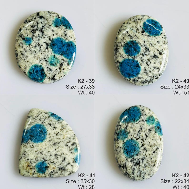 May include: Four polished K2 stone cabochons with white and grey granite-like speckled patterns and distinct bright blue circular inclusions. Each piece is labeled with a code, size in mm, and weight in carats: K2-39 (27x33mm, 40ct), K2-40 (24x33mm, 51ct), K2-41 (25x30mm, 28ct), and K2-42 (22x34mm, 40ct).
