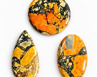 Könnte beinhalten: Drei polierte Cabochons mit einem brekziösen Muster aus leuchtend orangefarbenen, senfgelben, schwarzen und grauen Steinfragmenten. Das Set enthält einen runden, einen tropfenförmigen und einen ovalen Stein mit einer glatten Oberfläche.