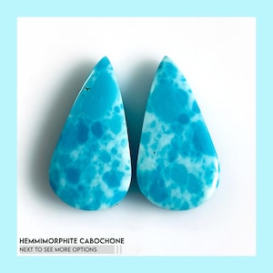 Puede incluir: Dos cabujones de hemimorfita en forma de lágrima con un vibrante patrón moteado en azul cielo y blanco con una superficie pulida y lisa. El texto en la parte inferior dice HEMIMORPHITE CABOCHONE NEXT TO SEE MORE OPTIONS.