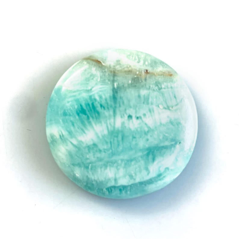 Peut inclure: Une pierre pr&eacute;cieuse polie de forme circulaire avec une surface lisse pr&eacute;sentant un motif marbr&eacute; aux couleurs turquoise, bleu sarcelle et blanc. La pierre affiche des stries naturelles fluides et une fine bande horizontale brun clair.