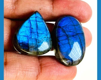 May include: Two natural blue labradorite gemstones. One is teardrop-shaped, the other is oval. Both stones have a vibrant blue iridescence with dark inclusions. The text "NATURAL BLUE LABRADORITE" and "NEXT TO SEE MORE OPTIONS" are visible.