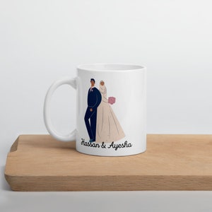 Op de afbeelding: Witte keramische mok met een zwart-witte illustratie van een bruid en bruidegom. De bruid draagt een witte jurk en een hoofddoek. De bruidegom draagt een zwart pak. De tekst "Hassan & Ayesha" staat op de mok gedrukt.