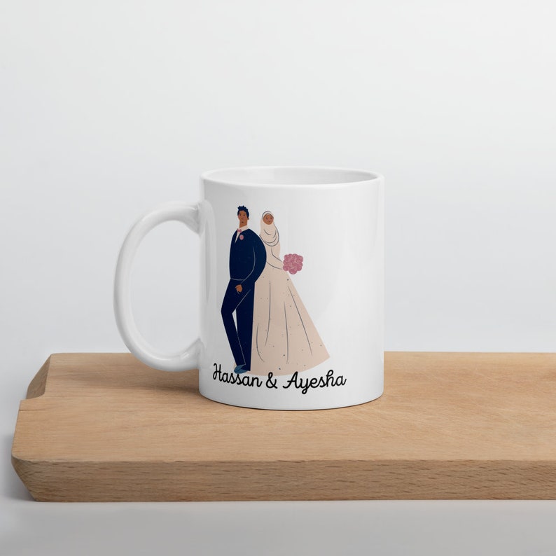 Op de afbeelding: Witte keramische mok met een zwart-witte illustratie van een bruid en bruidegom. De bruid draagt een witte jurk en een hoofddoek. De bruidegom draagt een zwart pak. De tekst "Hassan & Ayesha" staat op de mok gedrukt.