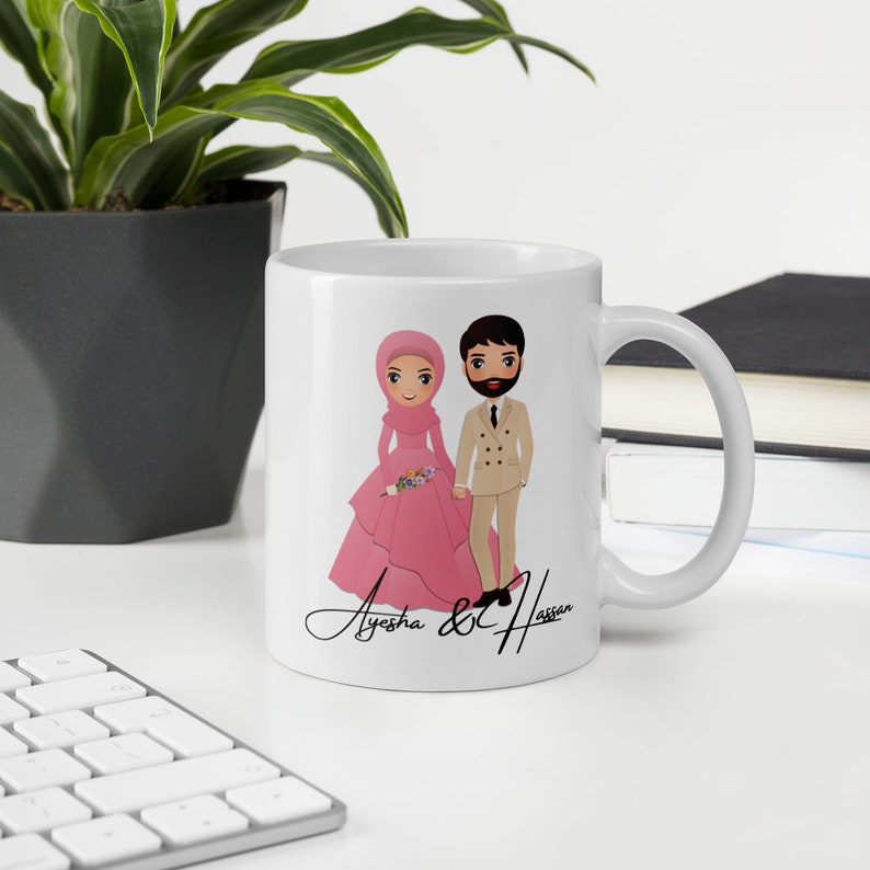 Op de afbeelding: Een witte keramische mok met een cartoonillustratie van een stel in trouwkledij. De vrouw draagt een roze hijab en jurk, en de man draagt een beige pak. De tekst "Ayesha & Hassan" staat in cursief onder het stel.