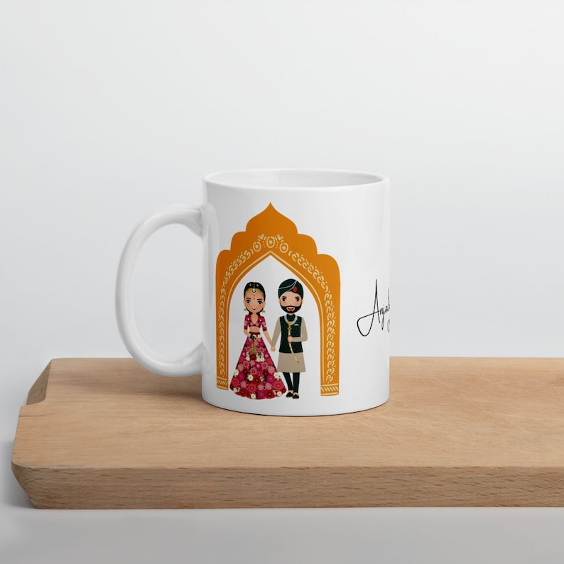 Puede incluir: Taza de cer&aacute;mica blanca con una ilustraci&oacute;n colorida de una pareja con atuendos tradicionales de boda india de pie bajo un arco dorado. El texto "Anjali &" est&aacute; escrito en cursiva en el lado derecho de la taza.
