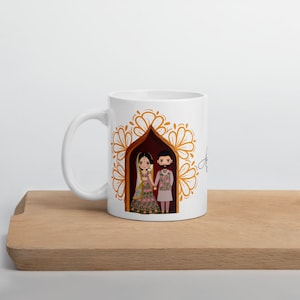 Puede incluir: Taza de cer&aacute;mica blanca con una ilustraci&oacute;n colorida de una pareja con atuendo tradicional de boda hind&uacute; de pie en una puerta decorada con dise&ntilde;os florales dorados.