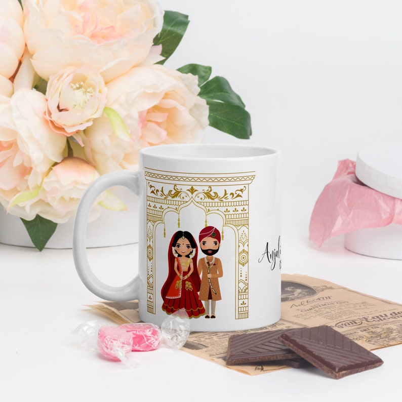 Puede incluir: Taza de cer&aacute;mica blanca con un dise&ntilde;o dorado de un arco de boda y una pareja de dibujos animados con atuendo tradicional de boda hind&uacute;. El texto "Anjali &" es visible en la taza.