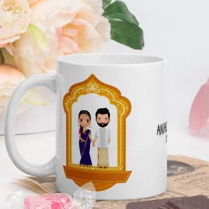Puede incluir: Taza de cer&aacute;mica blanca con una ilustraci&oacute;n dorada de una pareja con ropa tradicional india. La pareja est&aacute; de pie en una puerta con un marco dorado. El texto "ANUJ & SHIVANI" est&aacute; impreso en la taza.