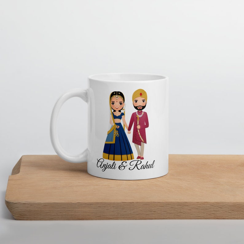 Puede incluir: Taza de cer&aacute;mica blanca con una ilustraci&oacute;n de dibujos animados de una novia y un novio con atuendos tradicionales de boda india. El texto "Anjali & Rahul" est&aacute; impreso debajo de la ilustraci&oacute;n.