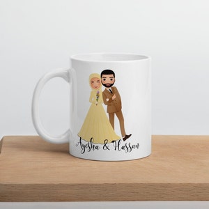 Op de afbeelding: Witte keramische mok met een cartoonillustratie van een bruid en bruidegom. De bruid draagt een gele jurk en een hoofddoek. De bruidegom draagt een bruin pak. De tekst "Ayesha & Hassan" staat op de mok gedrukt.