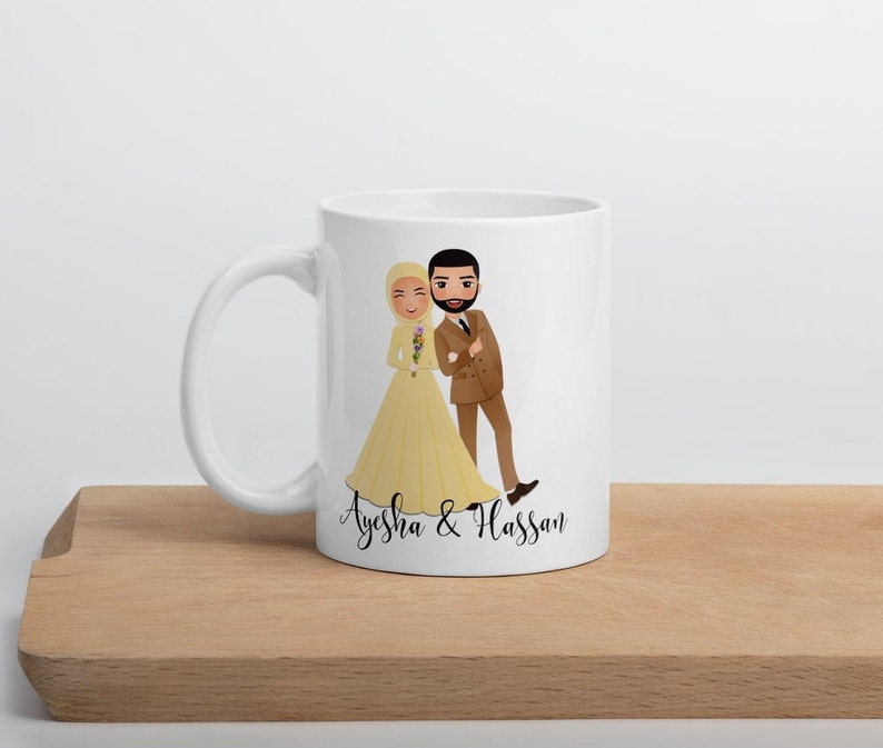Op de afbeelding: Witte keramische mok met een cartoonillustratie van een bruid en bruidegom. De bruid draagt een gele jurk en een hoofddoek. De bruidegom draagt een bruin pak. De tekst "Ayesha & Hassan" staat op de mok gedrukt.