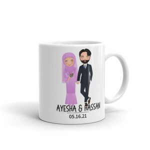 Op de afbeelding: Witte keramische mok met een cartoonillustratie van een stel in trouwtenue. De bruid draagt een roze hijab en jurk, en de bruidegom draagt een zwart pak. De tekst "AYESHA & HASSAN 05.16.21" staat op de mok gedrukt.
