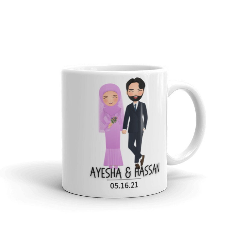 Op de afbeelding: Witte keramische mok met een cartoonillustratie van een stel in trouwtenue. De bruid draagt een roze hijab en jurk, en de bruidegom draagt een zwart pak. De tekst "AYESHA & HASSAN 05.16.21" staat op de mok gedrukt.