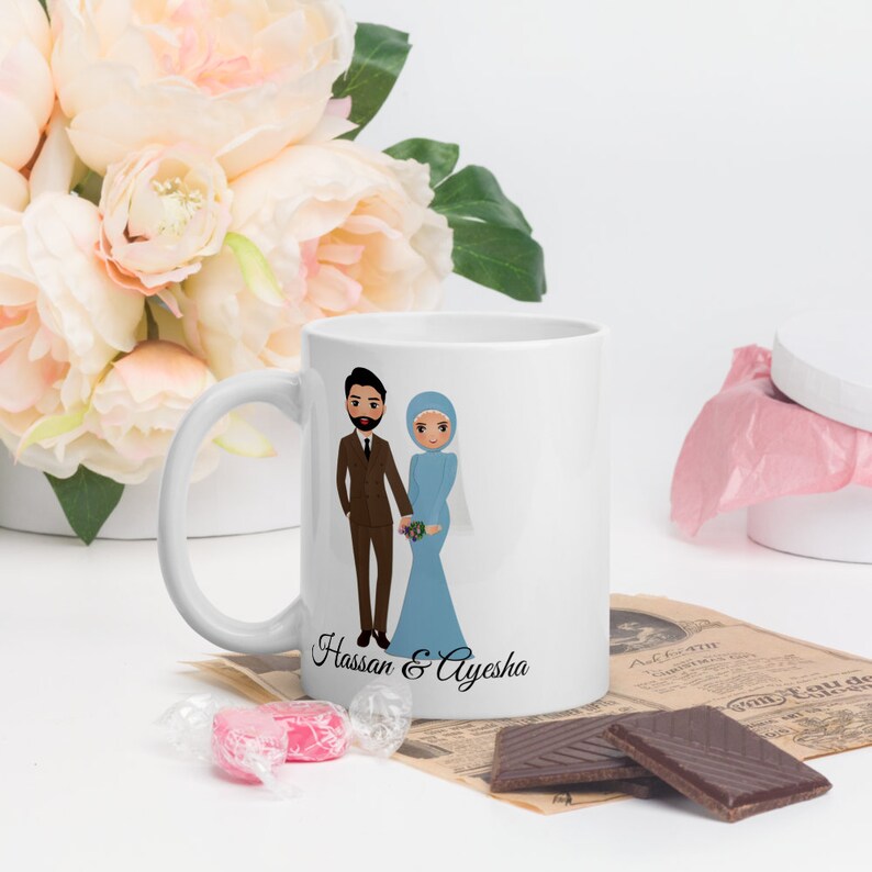 Op de afbeelding: Witte keramische mok met een cartoonillustratie van een stel in trouwtenue. De man draagt een bruin pak en de vrouw draagt een blauwe jurk en een hoofddoek. De tekst "Hassan & Ayesha" staat onder het stel gedrukt.