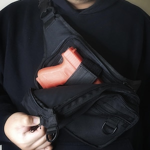 May include: Black tactical fanny pack with multiple compartments and a bright orange handgun inside. The pack has a zippered main compartment, adjustable straps, and a D-ring attachment. The person is wearing a black hoodie.