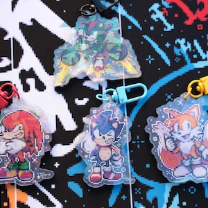 May include: A collection of clear acrylic keychains featuring pixelated images of Sonic the Hedgehog characters. Each keychain has a different colored clasp, including red, blue, and yellow. The characters include Sonic, Tails, Knuckles, and Shadow.