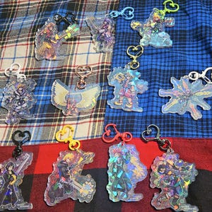 May include: A collection of 11 colorful, iridescent keychains featuring various characters from the Kingdom Hearts video game series. The keychains are made of clear plastic and have a holographic finish. Each keychain features a different character from the game, including Sora, Riku, Kairi, and others.