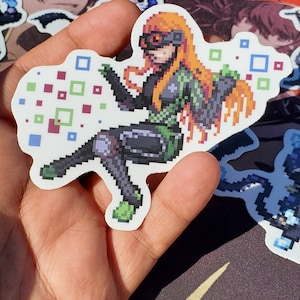 Pode incluir: Um adesivo de pixel art de uma personagem feminina com cabelo laranja, vestindo uma roupa verde e preta, sentada de pernas cruzadas e cercada por quadrados coloridos.