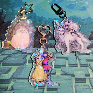 May include: Three iridescent keychains featuring characters from popular anime. The first keychain features Totoro, a fluffy, gray creature with large eyes. The second keychain features a white wolf with a girl riding on its back. The third keychain features two characters, a girl with long blonde hair and a boy with short brown hair, standing together.