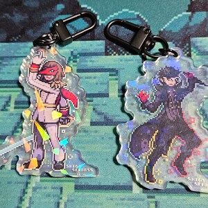 Peut inclure: Deux porte-clés irisés représentant des personnages pixélisés du jeu vidéo Persona 5. Le porte-clés de gauche représente un personnage portant une tenue rouge et blanche avec un masque. Le porte-clés de droite représente un personnage portant une tenue noire avec un masque. Les deux porte-clés portent l'inscription "SHEEP ST. BRASS".