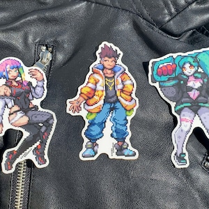 Könnte beinhalten: Drei Aufkleber im Pixel-Art-Stil mit Figuren mit verschiedenen Frisuren und Kleidung. Die Figuren sind eine Frau mit pinkfarbenem und blauem Haar, ein Mann mit braunem Haar und eine Frau mit grünem Haar. Die Aufkleber befinden sich auf einer schwarzen Lederjacke.
