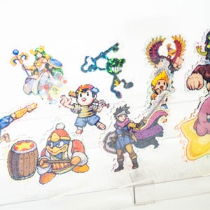 May include: A set of 10 colorful pixel art character stickers featuring characters from popular video games. The stickers are on a clear acrylic display stand.