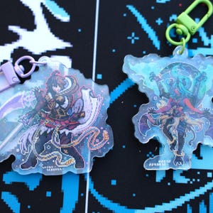 Könnte beinhalten: Zwei transparente Acryl-Schlüsselanhänger mit pixeliger Fantasy-Kunst. Einer zeigt eine Figur mit Schwert und dem Text "SHIDY SERBASS". Der andere zeigt eine mehrarmige Figur. Beide haben farbige Verschlüsse.