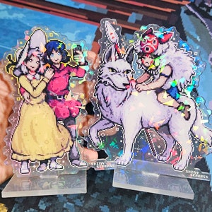 K&ouml;nnte beinhalten: Zwei schimmernde Acryl-St&auml;nder mit Anime-Charakteren. Der erste St&auml;nder zeigt ein M&auml;dchen mit langen blonden Haaren, das ein gelbes Kleid tr&auml;gt, und ein rothaariges M&auml;dchen, das ein rotes Kleid tr&auml;gt. Der zweite St&auml;nder zeigt ein M&auml;dchen mit kurzen braunen Haaren, das einen wei&szlig;en Wolf reitet. Beide St&auml;nder haben den Text "Shiny Seabass" auf der Unterseite.