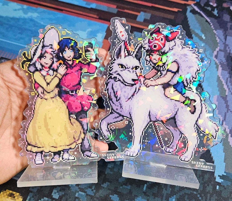 K&ouml;nnte beinhalten: Zwei schimmernde Acryl-St&auml;nder mit Anime-Charakteren. Der erste St&auml;nder zeigt ein M&auml;dchen mit langen blonden Haaren, das ein gelbes Kleid tr&auml;gt, und ein rothaariges M&auml;dchen, das ein rotes Kleid tr&auml;gt. Der zweite St&auml;nder zeigt ein M&auml;dchen mit kurzen braunen Haaren, das einen wei&szlig;en Wolf reitet. Beide St&auml;nder haben den Text "Shiny Seabass" auf der Unterseite.