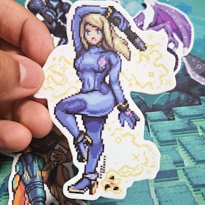 May include: A pixelated sticker featuring a character in a blue bodysuit, holding a weapon. The sticker has a white border and a yellow energy effect. The character has blonde hair and is in a dynamic pose. The sticker is held by a hand.
