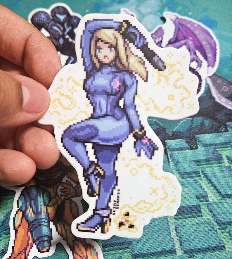 May include: A pixelated sticker featuring a character in a blue bodysuit, holding a weapon. The sticker has a white border and a yellow energy effect. The character has blonde hair and is in a dynamic pose. The sticker is held by a hand.