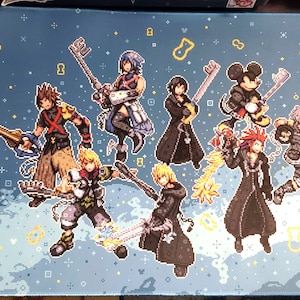 May include: A blue and yellow gaming mouse pad featuring a pixelated design of characters from the Kingdom Hearts video game series. The characters include Sora, Riku, Kairi, Mickey Mouse, Donald Duck, Goofy, and others. The mouse pad has a blue background with yellow stars and keyhole symbols.