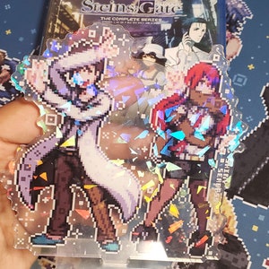 May include: A holographic acrylic stand featuring pixelated characters from the anime series "Steins;Gate". The stand features two characters, one in a white lab coat and the other in a red jacket. The stand is on a clear acrylic base.