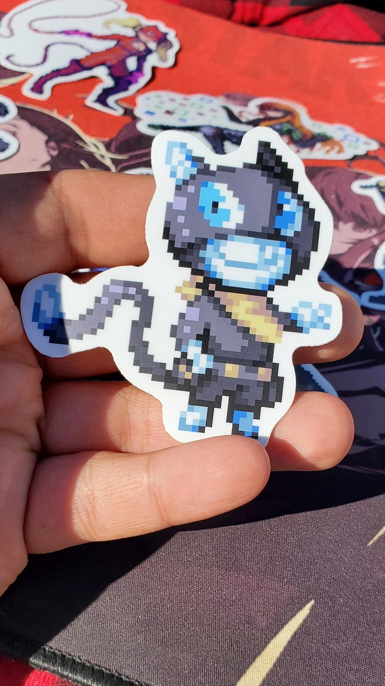 Pode incluir: Um adesivo de arte de pixel preto e azul de uma criatura semelhante a um gato com um cachecol amarelo. A criatura tem um rosto azul com olhos brancos e um largo sorriso.