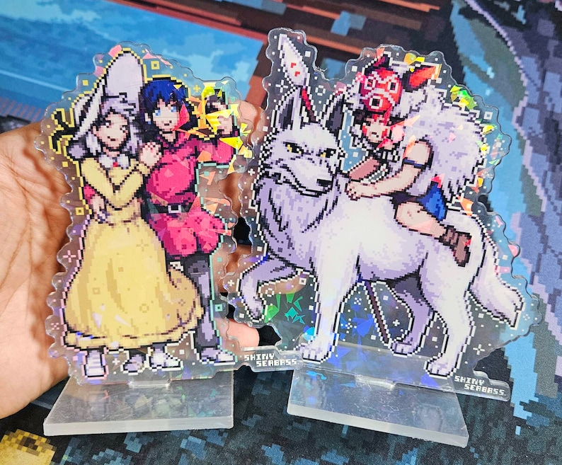 K&ouml;nnte beinhalten: Zwei Acryl-Charakter-St&auml;nder mit Anime-Charakteren. Der erste St&auml;nder zeigt zwei weibliche Charaktere, eine mit einem gelben Kleid und einem wei&szlig;en Hut und die andere mit einem roten Kleid. Der zweite St&auml;nder zeigt einen wei&szlig;en Wolf mit einem Charakter, der auf seinem R&uuml;cken reitet. Beide St&auml;nder haben eine glitzernde, irisierende Oberfl&auml;che und den Text "Shiny Seabass" auf der Unterseite.