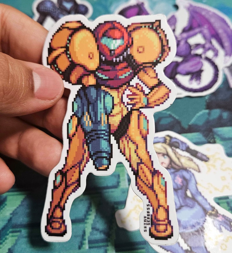 May include: A pixelated sticker of Samus Aran from the Metroid series. The sticker features the character in her iconic orange and yellow power suit, with a teal arm cannon. Other stickers are visible in the background.