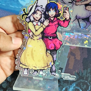 K&ouml;nnte beinhalten: Ein pixeliger Acryl-St&auml;nder mit zwei Figuren, einem M&auml;dchen mit wei&szlig;en Haaren und einem gelben Kleid und einem Jungen mit blauen Haaren und einem roten Hemd. Der Junge h&auml;lt ein Telefon und das M&auml;dchen h&auml;lt seinen Arm. Der St&auml;nder hat eine holografische Oberfl&auml;che und der Text "Shiny Seabass" ist unten aufgedruckt.