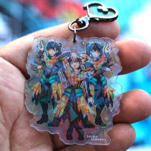 May include: A clear, pixelated keychain featuring three anime-style figures in colorful outfits. The figures are posed with their hands up. The keychain has a silver clasp and the text "SHINY SERBASS".