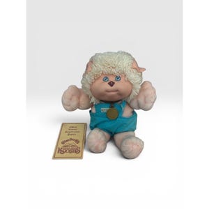 Puede incluir: Un peluche Cabbage Patch Kids Koosas con cabello de hilo color crema, ojos azules y nariz marrón. El peluche viste un overol de pana color verde azulado con un collar marrón y un medallón dorado. Una etiqueta rectangular muestra el texto Koosas.
