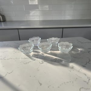 May include: A set of five small, clear glass bowls with a scalloped edge design. Each bowl features a rounded base and a textured, petal-like rim, reflecting light across the surface. These transparent glass vessels are arranged in a cluster.
