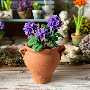 Peut inclure: Un pot en terre cuite à deux anses, rempli d'une petite composition florale violette. Le pot a un motif décoratif sur le devant.