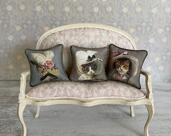 May include: Three decorative throw pillows with a vintage aesthetic feature embroidered images of cats wearing hats. The pillows are arranged on a white, ornate sofa with a floral patterned fabric.
