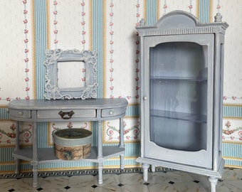 May include: A gray wooden console table with two drawers and a shelf, and a gray wooden cabinet with glass doors and shelves. The furniture is in a room with floral wallpaper and a black and white tiled floor.